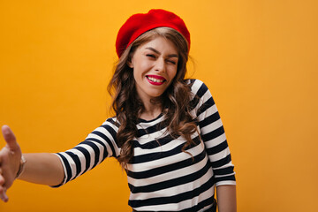 Woman in striped shirt and beret winking and taking selfie on orange background. Attractive girl in fashionable outfit posing..