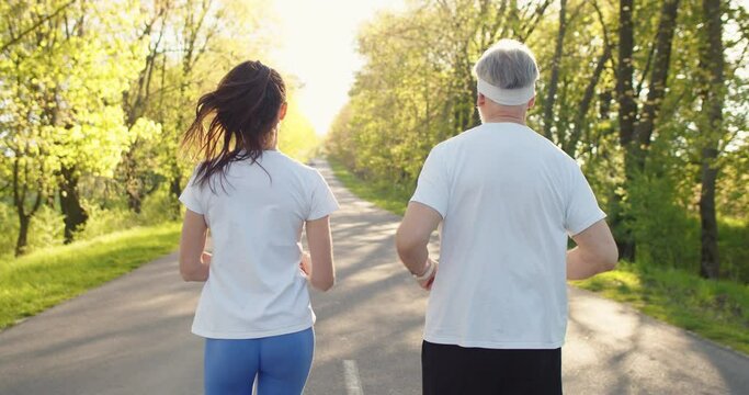 Back view of Caucasian old grey-haired man running outdoors exercising with young woman. Father and daughter jogging on street outside. Sport lifestyle, fitness, working out together concept