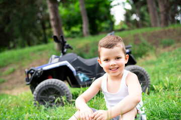 Little five-year-old child boy sit on the grass near his black and purple ATV.