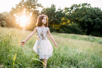 Playful caucasian child in summer dress walking among high grass on meadow. Pretty kid with brown hair enjoying sunrise on fresh air.