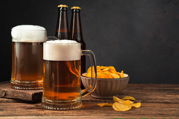 Two glasses of frothy beer, chips and beer bottles on a brown background.