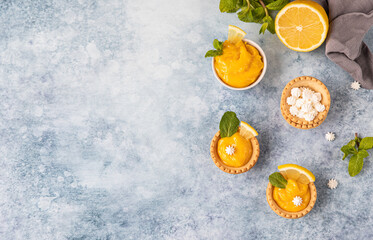 Mini tarts with lemon curd, mini meringue, lemon slices and mint on blue concrete background. Top view. Copy space.