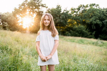 Pretty young girl wearing stylish white dress posing at green garden. Beautiful summer sunrise on background. Concept of childhood and carefree.