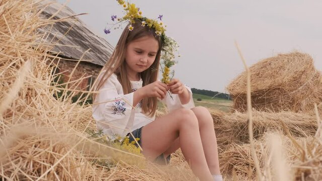 Ukrainian preteen girl wearing national dress sitting on hay stack and weave wreath with flowers. Pretty child in countryside in summertime. Female teen on farm with old house on background