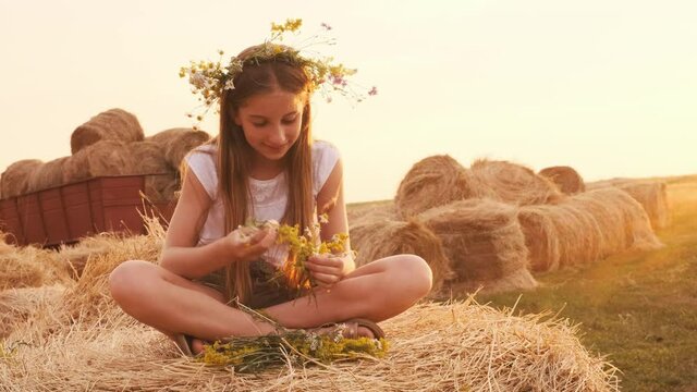 Ukrainian teen girl sitting on hay stack in the field and weave wreath with flowers. Pretty female teenager in countryside in summertime