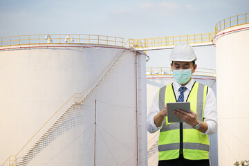 inspection engineer holding tablet computer against fuel tanks at industry site.