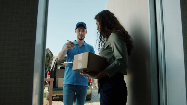 Female customer is opening house front door taking cardboard box from delivery worker signing paper getting order. People and postal service concept.