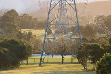 Base of large overland powerline tower on sunset with bushfire smoke in the background and green cricket oval with large eucalyptus trees surrounding.