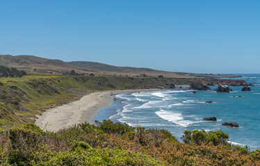 San Simeon, CA, USA - June 8, 2021: Pacific Ocean coastline. Closeup of beige sand beach whereon blue oceans crashes white surf under blue sky. Green covered cliffs form half circle.