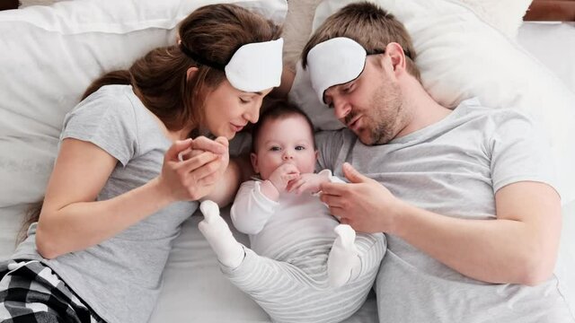 Beautiful family wearing same pajamas and sleeping eye masks lying in the bed with little child boy and petting him. Young parents with their toddler baby in bedroom. Father kisses hand of his kid