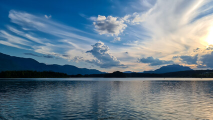 A panoramic view on the Lake Faak in Austria. The lake is surrounded by high Alpine peaks. The sun in slowly setting behind the mountains. Lots of clouds. Calm surface reflects the sunbeams. Serenity