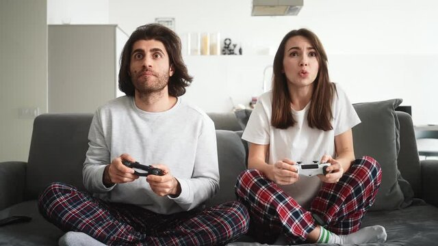 An emotional couple woman and man are playing console games sitting on the sofa indoors. Man is doing winner gesture