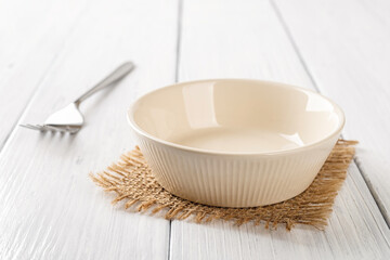 Fototapeta na wymiar Empty beige bowl and metal fork on a white wooden table. Clean porcelain soup bowl on a table. Empty ceramic tableware ramekin for food design mockup. High key image.