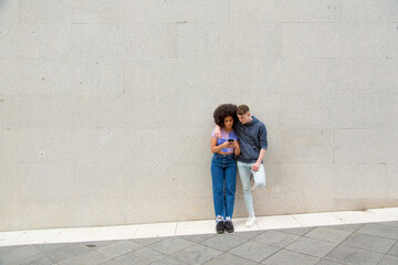 Plakat relationship friends young white man and black woman using mobile phone in the city with a background wall with copy space