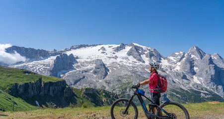 nice and active senior woman riding her electric mountain bike on the Pralongia Plateau in the Alta Badia Dolomites with glacier of Marmolata summit in Background, South Tirol and Trentino, Italy
