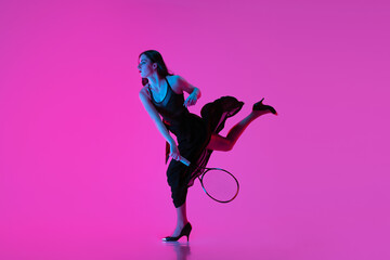 Fototapeta na wymiar High-fashion styled young woman in black evening gown, dress playing tennis isolated over pink neon background. Concept of fashion and sport