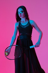 High-fashion styled young woman in black evening gown, dress playing tennis isolated over pink neon background. Concept of fashion and sport