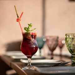 Red berries drink with mint sprig on sunny blurred background. Glass of refreshing drink with straws. Healthy fresh juice. Bright colors of nature. Copy space.