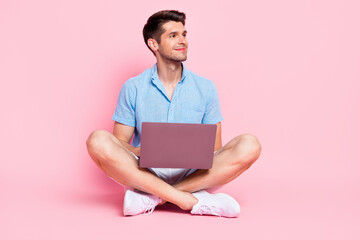 Full length body size photo smiling man in blue shirt sitting working on laptop looking copyspace isolated pastel pink color background