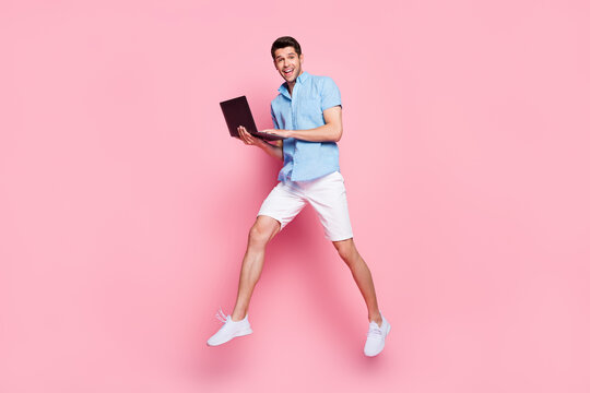 Photo of positive carefree man jump typing notebook run wear blue shirt shorts isolated on pink color background