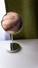 Figure of a fat woman is reflected in a round mirror. Mirror stands on a white table on green curtains background
