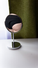 Figure of a fat woman is reflected in a round mirror. Mirror stands on a white table on green curtains background