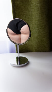 Figure of a fat woman is reflected in a round mirror. Mirror stands on a white table on green curtains background