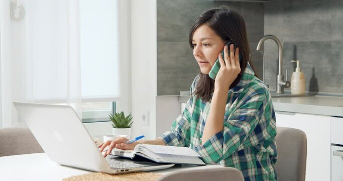 Young woman talking on cell phone working on laptop in home office. Female worker providing professional advice to a client remotely using wireless devices. Distant work.