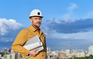 Construction site or building concept. Caucasian building contractor or engineer in white hardhat and orange jacket with laptop and blueprints in sunny day with blue cloudy sky and urban skyline