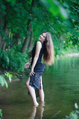 A beautiful young romantic girl with long flowing hair in a black long dress stands in the green water of a summer river or lake.