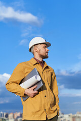 Architect, engineer on construction site or building concept. Building contractor man in white hardhat and orange jacket with laptop and blueprints in sunny day with blue cloudy sky and urban skyline