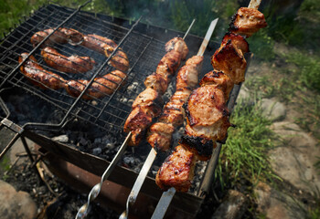 Barbeque meat and sausages or bratwurst on a grill grate in backyard. Man preparing shashlik or shish kebab over charcoal. Grilled meat on metal skewer outdoor. BBQ party or picnic food. Close up shot
