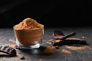 Dry carob pods and carob powder on a dark background. Organic healthy ingredient for vegan vegetarian food and drinks, close up - obrazy, fototapety, plakaty