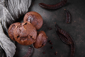 Chocolate carob cupcakes cooking with carob powder and dried carob pods on a dark background. Organic healthy sweet food for vegan vegetarian food and drinks, close up, above - obrazy, fototapety, plakaty