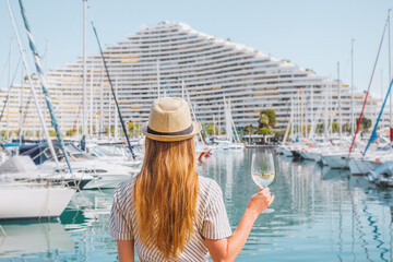 Blonde hair woman in a hat standing with her back on sea, yachts, buildings and boats background. White wine glass in hand. Vacation in Europe. Nice, French riviera. Travel photo. - obrazy, fototapety, plakaty