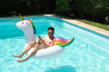 Young man wearing sunglasses using the phone and looking at a side on a unicorn inflatable ring in...