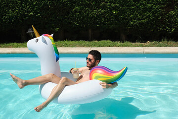 Young man wearing sunglasses drinking a refreshment on a unicorn inflatable ring in a swimming...