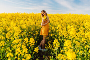 Naklejka na ściany i meble Attractive cheerful woman on rapeseed field with sunset light. Yellow flowers and caucasian woman
