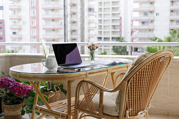 Home office concept. Designated work from home area at the balcony. Modern laptop, notebook and a cup of coffee on table, stylish straw chair with cushions. Close up, copy space, background.