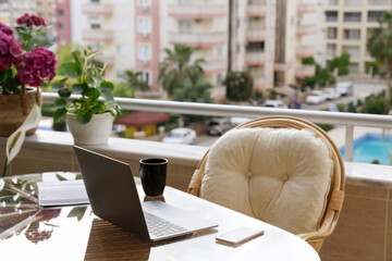 Home office concept. Designated work from home area at the balcony. Modern laptop, notebook and a cup of coffee on table, stylish straw chair with cushions. Close up, copy space, background.