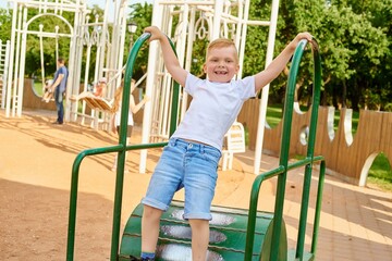 A small red-haired five-year-old boy is playing on the playground in the park in the summer