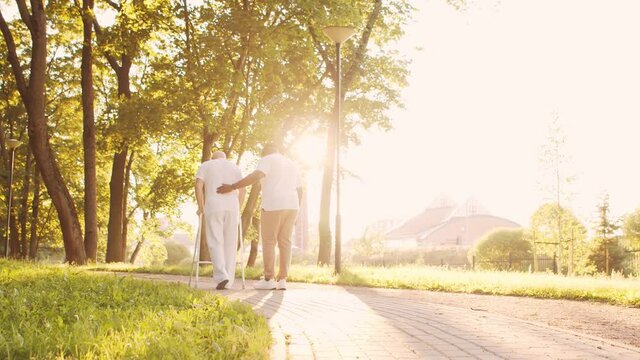 African-American caregiver is teaching disabled old man to walk with walker. Professional nurse and handicapped patient in the park. Assistance, rehabilitation and health care. Golden hour sunset.