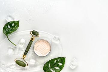 Cream, green jade face roller on ice with ice cubes and exotic leaves. Monstera Adansonii leaves on off white background. Beauty facial massage with home made moisturizer flat lay, copy-space. © tilialucida