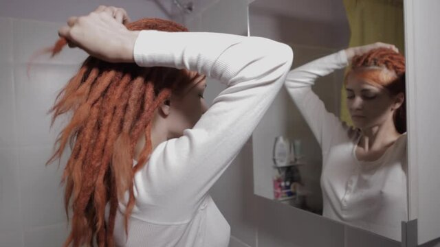 A young woman with red dreadlocks looks in the mirror and Ties dreadlocks, in bathroom in the morning getting ready during everyday routine at home