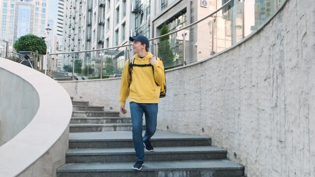Happy young courier with yellow uniform and backpack actively dancing while walking down the stairs. Successful delivery man concept