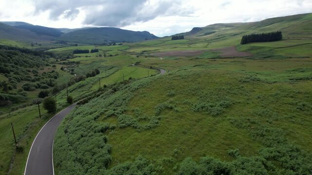 Aerial footage over a river flowing through a partially wooded glen with a long and winding rural road disappearing into the distance.