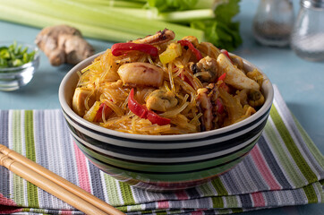 Funchoza with seafood and vegetables in a bowl on light blue background. Delicious Asian homemade food. Close-up