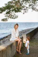 Young woman and toddler girl on sea vacation. Family sits on a beach at sunny day, enjoying summer holidays. Summer lifestyle portrait of pretty family: mom and daughter. Travel local tourism