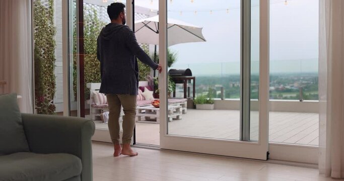 man opens sliding doors, going out on rooftop patio with cozy lounge zone and beautiful landscape view