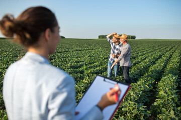 Female agronomist in foreground taking notes writing and two male farmers standing in soy field...